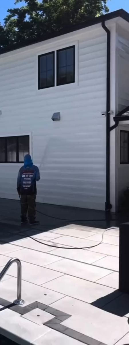 A technician washing the white siding at the back of a house beside a pool patio