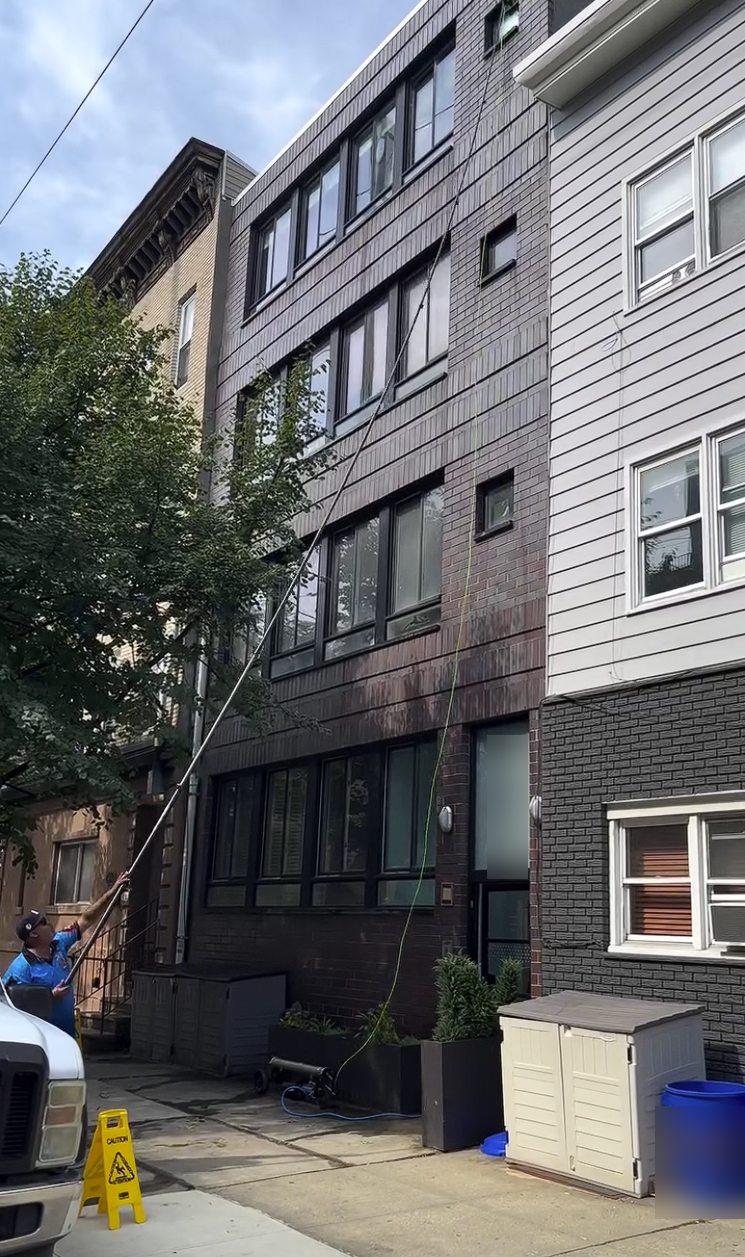 A Premier Power Washerz technician cleaning fourth floor windows on a Hoboken brick condo building with a water fed pole from the sidewalk