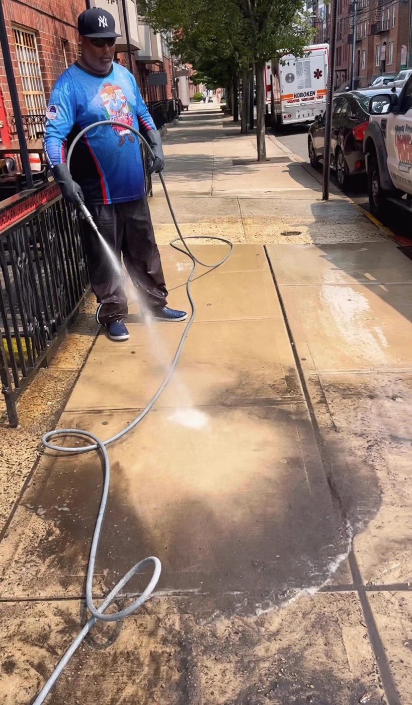 A technician rinsing the sidewalk with the cleaned section bright and the remaining grime visible ahead