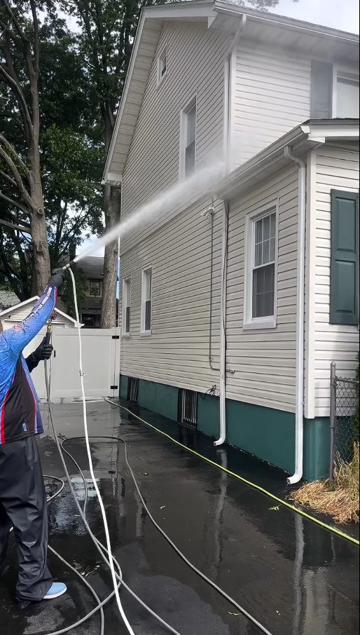 A technician soft washing the tall side elevation of the Irvington colonial above its green painted foundation